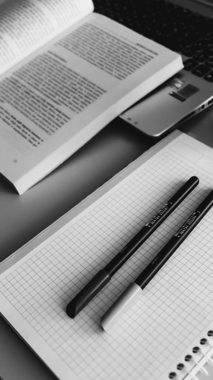 digital-01 Black and white closeup of a study desk with an open book, grid notebook, and pens for a minimalist study scene.